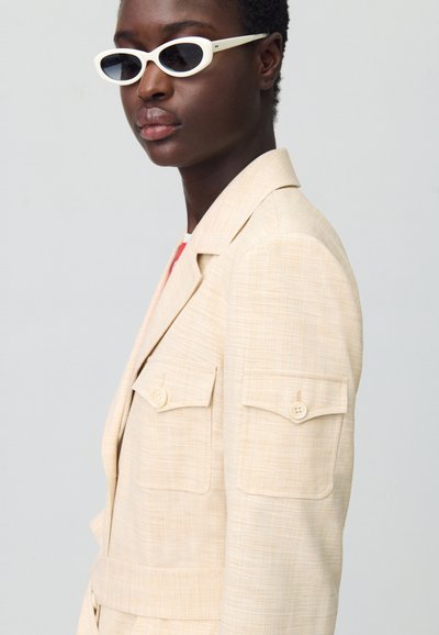 Blazer beige ajusté avec deux poches poitrine et accents de boutons, accompagné de lunettes de soleil ovales blanches. Tissu lisse avec une texture subtile.