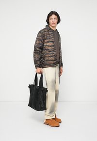 Jonge man in een jas met camouflagepatroon, lichte aflopende broek, bruine laarzen, die een grote zwarte tote bag vasthoudt, staand tegen een witte achtergrond.