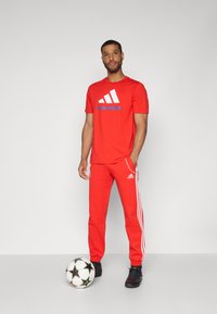 Rød trenings T-skjorte med hvite og blå logoer, kombinert med røde joggebukser med hvite striper og merke. Svarte joggesko og en fotball.