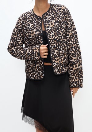 Femme portant une veste boutonnée imprimé léopard sur un crop top noir et une jupe noire asymétrique avec bordure en dentelle, debout devant un fond blanc.