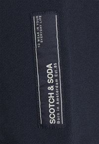Navy Stoffetikett mit dem weißen Logo "SCOTCH & SODA", sowie den Texten "Born in Amsterdam Est. 85" und "To Wear in Your Everyday Life."