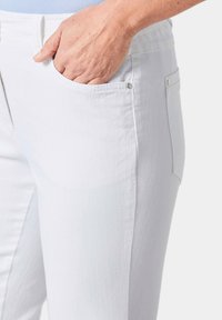 Weiße Denim-Hose mit glatter Textur, ausgestattet mit einer Seitentasche und dekorativem Druckknopf-Detail am Taillenbund.