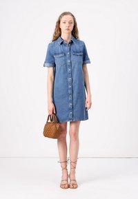 Denim skjortekjole, kne-lengde, blå med korte oppbrettede ermer, to brystlommer, frontlukking med knapper, kombinert med en liten brun veske.