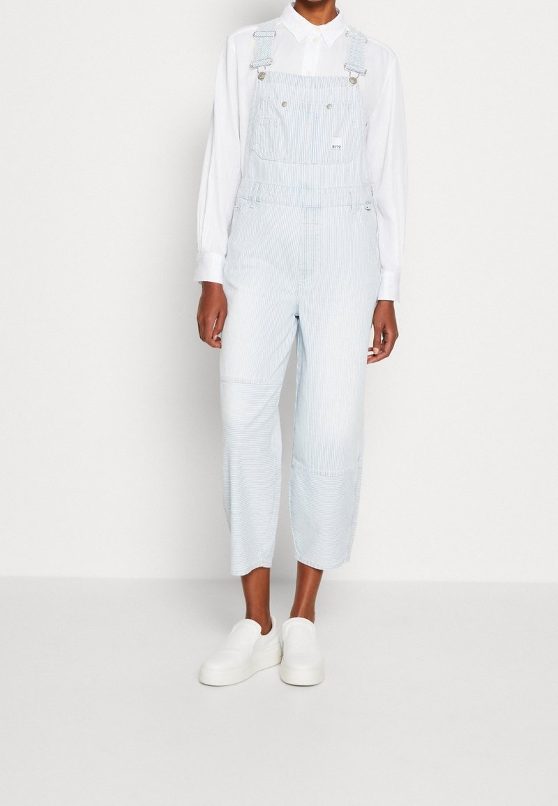 Personne portant une salopette rayée bleu clair et blanc par-dessus une chemise blanche à manches longues avec col, et des chaussures blanches sans lacets, debout.