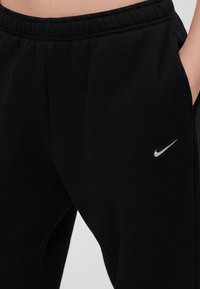 Μαύρη φόρμα Nike με ελαστική μέση και λευκό λογότυπο Swoosh στο δεξί μηρό, το χέρι τοποθετημένο στην δεξιά τσέπη.