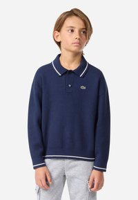 Σκούρο μπλε πλεχτό μπλουζάκι με άσπρη ριγέ κολάρο, διαθέτοντας τρεις κουμπιά και ένα μικρό κεντημένο λογότυπο στο στήθος.