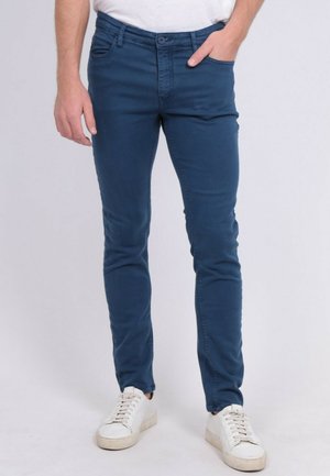 Homme portant un pantalon bleu marine cintré et des baskets blanches, debout avec une main dans la poche devant un fond uni.