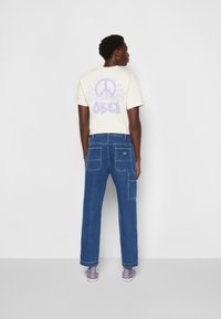 Cream graphic t-shirt featuring purple "OBEY" design, paired with blue wide-leg jeans and gray sneakers. Visible back pockets and stitching details.