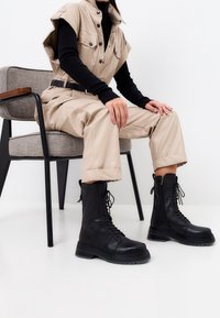 Stivali da combattimento neri con lacci e suola spessa, con una finitura in pelle liscia e cerniera laterale, indossati con pantaloni cargo beige e una maglietta nera.