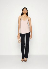 Mujer con un top de satén rosa claro, pantalones negros, sandalias negras, sosteniendo un bolso texturizado beige contra un fondo blanco.