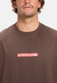Brun bomulds t-shirt med rund hals, der har et rødt rektangulært logo "QUIKSILVER" i hvid tekst på forsiden.