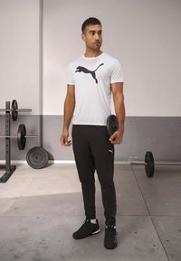 Witte sport t-shirt met zwart Puma-logo, zwarte slim-fit sportbroek en zwarte sneakers. Sportschoolomgeving met gewichten op de achtergrond.