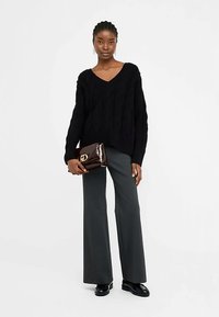 Pulover negru tricotat cu model în relief, cu decolteu în V, asortat cu pantaloni gri evazați și mocasini negri. Modelul ține o poșetă plic maro închis cu un logo.