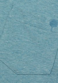Tessuto azzurro chiaro e testurizzato con una tasca a toppa decorata da un design di albero ricamato e macchie di blu scuro in tutto il tessuto.