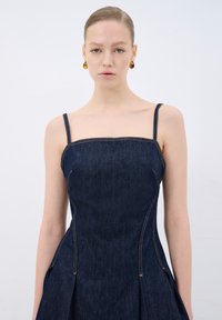 Denimdressi, jossa on ohuet hihnat, jämäkkä yläosa ja levenevä helma. Ominaisuuksina oranssitommennukset ja syvä indigo väri. Sujuva rakenne.