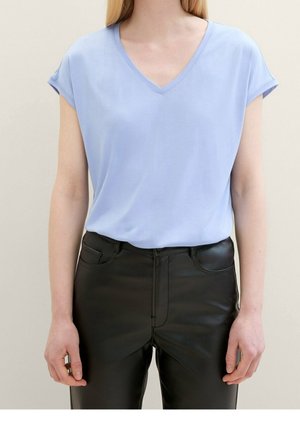 Femme portant un haut bleu clair à col en V manches courtes et un pantalon en cuir noir taille haute, debout devant un fond uni.