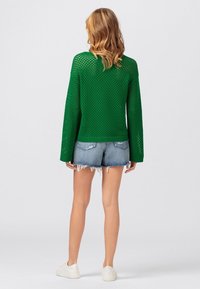 Kvinna med vågigt blont hår som bär en grön öppenstickad tröja, slitna denimshorts och vita sneakers, stående med ryggen mot kameran på en enkel bakgrund.