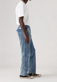 Homme debout de profil portant un t-shirt blanc, un jean en denim bleu clair avec un rabat de poche arrière boutonné, et des baskets noires.