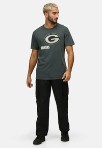 Šedé bavlněné tričko se velkým oválným logem s písmenem "G" a nápisem "PACKERS". Kombinováno s černými cargo kalhotami a černými botami.