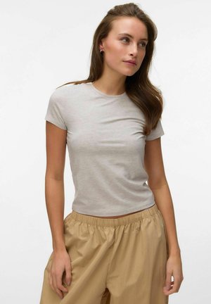 Kvinde med langt brunt hår iført en tætsiddende lys grå t-shirt med korte ærmer og løse beige bukser, stående mod en hvid baggrund.