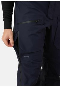 Marineblauwe waterdichte broek met meerdere gecremete ritszakken, versterkte naden, en een Recco®-logo. De stof heeft een gladde, duurzame textuur.