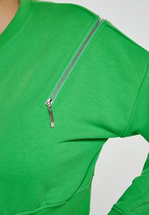 Zielona bluza z prążkowanym dekoltem i srebrnym zamkiem na lewym ramieniu, wykonana z gładkiego, lekkiego materiału.