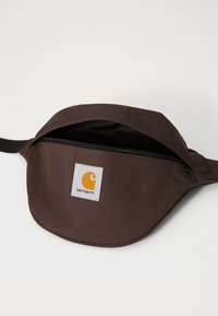 Braune Stoffumhängetasche mit einem Reißverschluss, ausgestattet mit einem weißen Etikett mit gelbem Logo. Glatte Textur und geschwungenes Design. Verstellbarer Riemen.