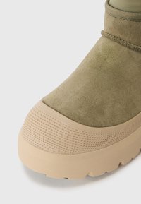 Olivgrön mockabot med en beige texturerad gummisula, rund tå och förstärkt tåhätta. Har en sydd söm vid vristen.