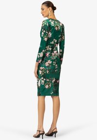 Abito verde in velluto con maniche lunghe, motivo floreale nei toni del rosa e bianco, silhouette aderente, lunghezza al ginocchio, abbinato a tacchi neri.