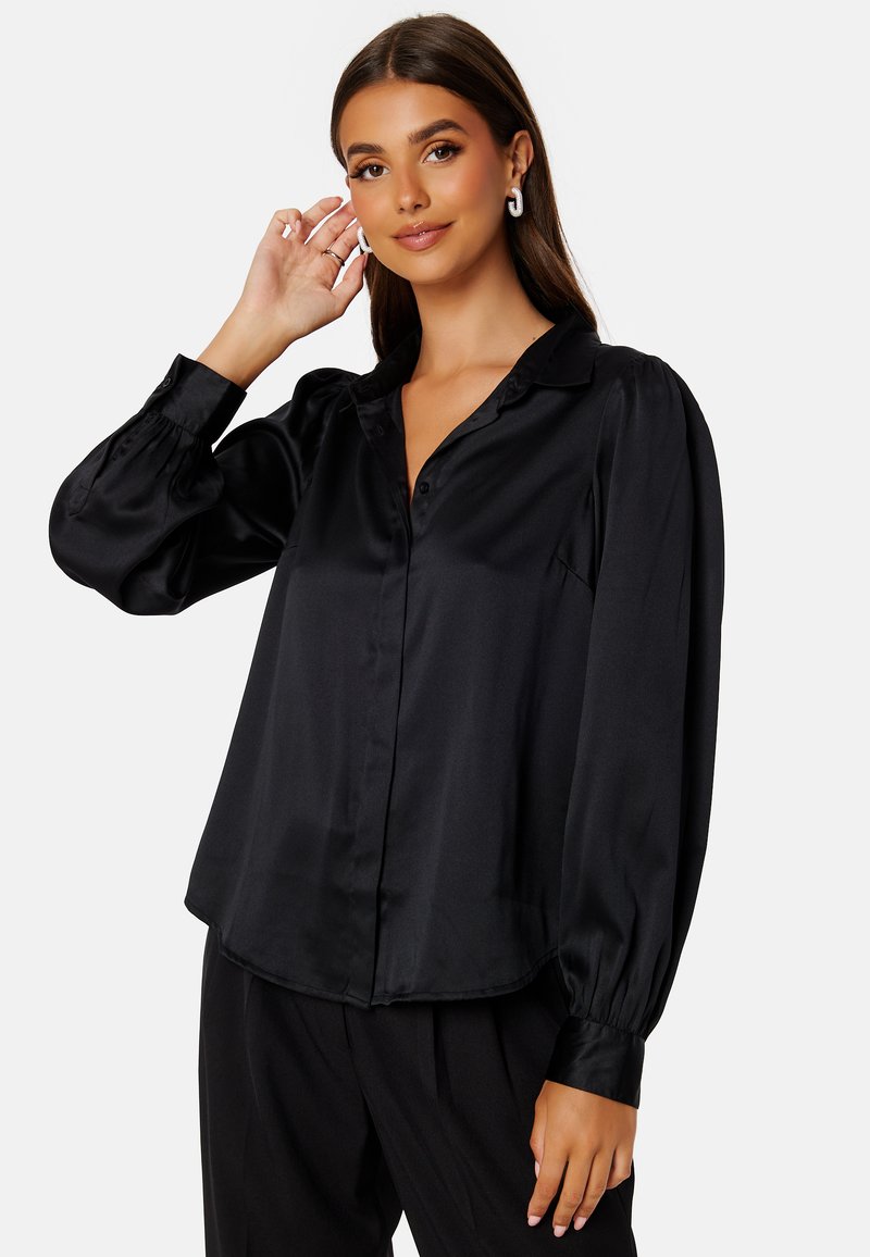 Blusa nera in satin con colletto, chiusura a bottone frontale e maniche lunghe a sbuffo. Tessuto liscio con una vestibilità leggermente ampia e una leggera lucentezza.