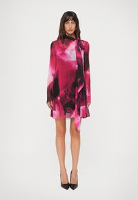 Mulher em pé, vestindo um vestido longo de manga longa com padrões florais em rosa vibrante e preto, com uma gravata no pescoço, combinado com sapatos de salto alto pretos e pontiagudos.