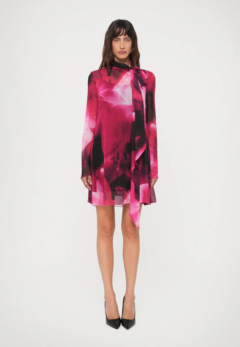 Mulher em pé, vestindo um vestido longo de manga longa com padrões florais em rosa vibrante e preto, com uma gravata no pescoço, combinado com sapatos de salto alto pretos e pontiagudos.