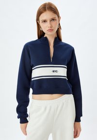 Sudadera recortada de color azul marino con un cuello alto, cierre con cremallera y franjas horizontales blancas que presentan el logo "NYC" centrado en el pecho. Diseño minimalista.