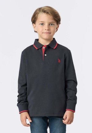 Ragazzo con capelli castano chiaro che indossa una polo a maniche lunghe blu navy con rifiniture rosse e jeans blu, in piedi davanti a uno sfondo bianco uniforme.