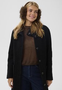 Juodas tekstūruotas paltas su dideliais mygtukais, po juo rudas megztinis, rudi kailiniai ausų apsaugai. Modelis pozuoja neutraliame fone.