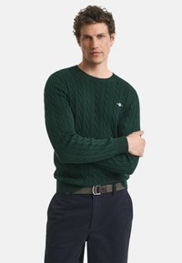Dunkelgrüner Kabelstrickpullover mit rundem Halsausschnitt und gerippten Bündchen, getragen mit marineblauen Hosen und einem schwarzen Gürtel mit einer silbernen Schnalle.