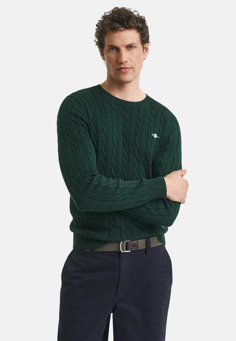 Dunkelgrüner Kabelstrickpullover mit rundem Halsausschnitt und gerippten Bündchen, getragen mit marineblauen Hosen und einem schwarzen Gürtel mit einer silbernen Schnalle.