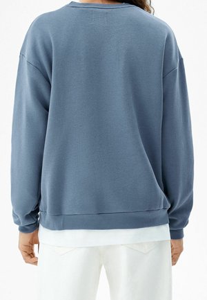 Person trägt einen lockeren blauen Pullover über einem weißen Hemd und weißen Hosen, von hinten vor einem einfarbigen Hintergrund gesehen.