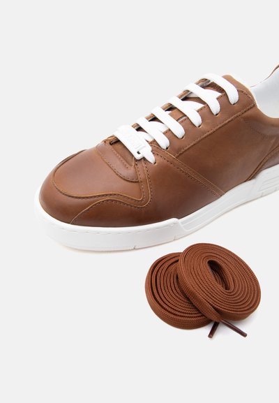 Καφέ δερμάτινο αθλητικό παπούτσι με λευκή σόλα και κορδόνια, συνοδευόμενο από δύο καφέ κορδόνια τυλιγμένα σε πηνία, σε λευκό φόντο.