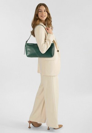 Femme en tailleur beige et talons tenant un sac à bandoulière vert foncé, souriante, debout devant un fond clair uni.