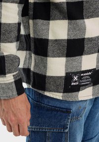 Camisa de franela a cuadros en blanco y negro con una etiqueta, mostrando una textura de lana y un lado con cremallera. Combinada con pantalones de mezclilla azules.