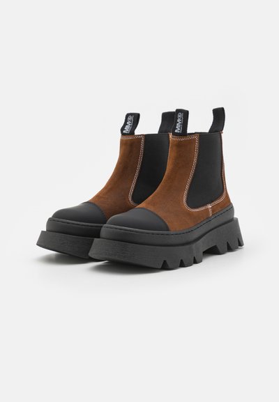 Botas de tobillo de ante marrón con paneles laterales elásticos negros y suelas de goma negra gruesas que presentan un acabado de borde en zigzag.