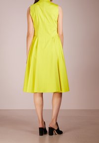 Bezramenné šaty v jasně žluté barvě s upnutým korzetem a rozšířenou sukní, vyrobené z hladkého materiálu. Černé lodičky na podpatku celý vzhled doplňují.