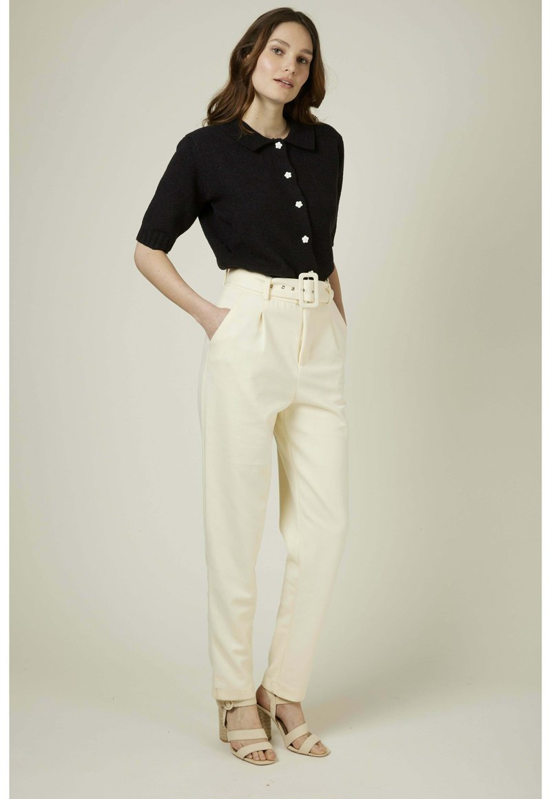 Pull noir à manches courtes avec un col et des boutons en forme de fleur, associé à un pantalon crème taille haute avec une large ceinture et des plis.
