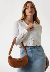 Sac à main en simili cuir marron avec une forme incurvée, matériel en acier inoxydable et détails cousus. Le sac est équipé d'une bandoulière et d'un accent logo.