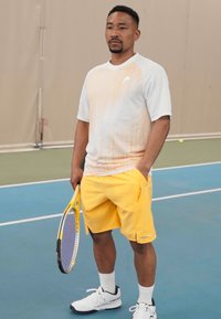 Tennisspieler in einem leichten, kurzärmligen, weißen und orangefarbenen Hemd mit Farbverlauf und gelben Shorts, der einen Tennisschläger hält.