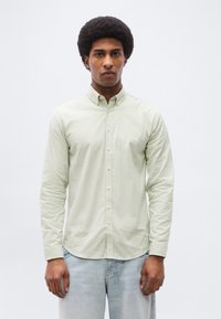 Άνδρας με κατσαρά μαλλιά, φορώντας πουκάμισο με κάθετες ρίγες σε ανοιχτό πράσινο και λευκό και ανοιχτό μπλε τζιν, στέκεται μπροστά σε λευκό φόντο.