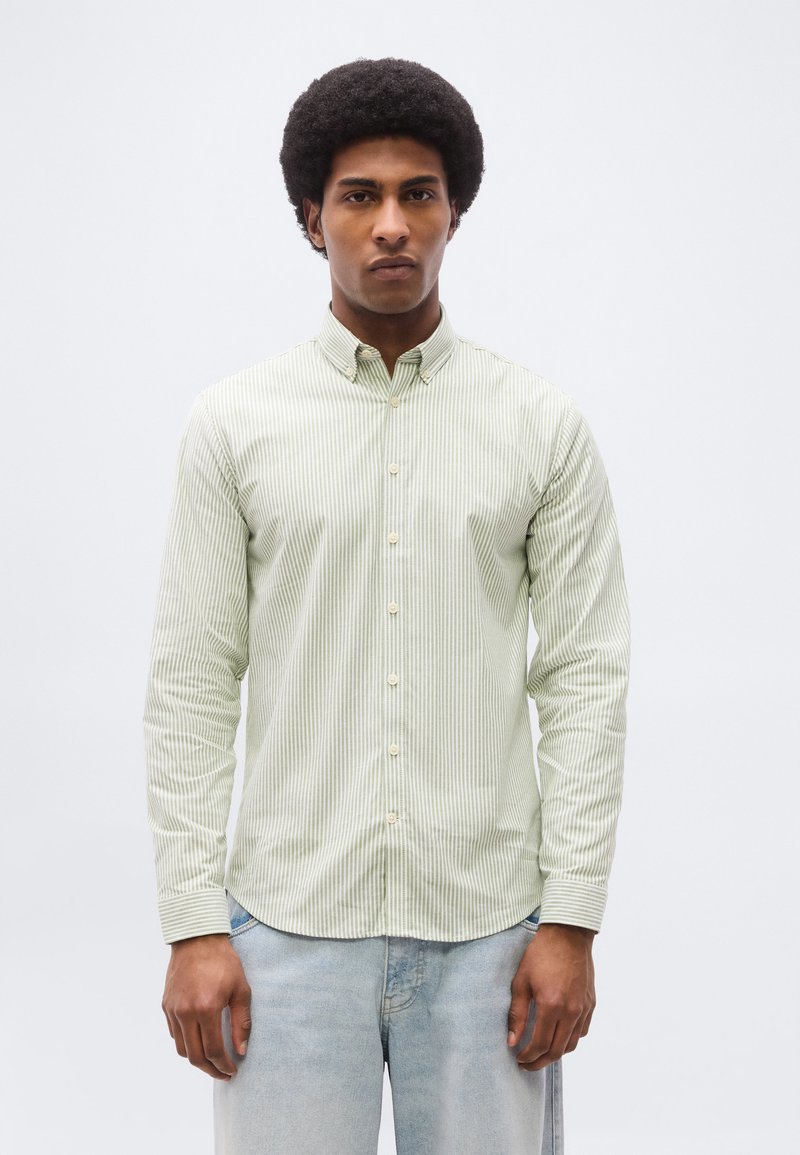 Άνδρας με κατσαρά μαλλιά, φορώντας πουκάμισο με κάθετες ρίγες σε ανοιχτό πράσινο και λευκό και ανοιχτό μπλε τζιν, στέκεται μπροστά σε λευκό φόντο.