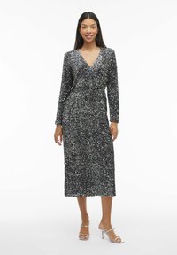 Abito lungo di paillettes grigio scuro con scollatura a V e maniche lunghe, caratterizzato da una vita aderente e una lunghezza fino al polpaccio. Abbinato a tacchi alti argentati.