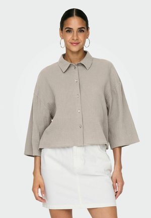 Camiseta cortada de color gris claro con cuello tipo polo, botones en el frente y mangas anchas y sueltas. Tejido suave y texturizado con un patrón sutil.